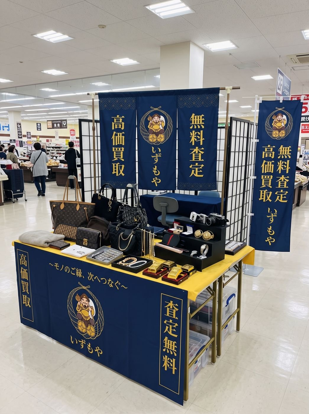 スーパーの店内に設営されたいずもやの催事ブース。紺地に金文字ののれんと巻き幕、黄色のクロスを掛けた陳列机にバッグや時計、貴金属が並ぶ